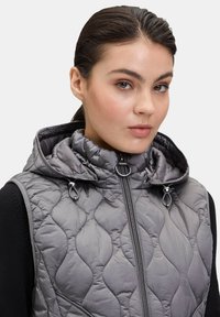 Dżinsowa szara kamizelka z wysokim kołnierzem i zamkiem błyskawicznym, z regulacją w postaci sprzętu i teksturowaną powierzchnią, zaprojektowana dla ciepła i do noszenia warstwowego.