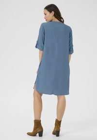 Abito camicia blu con vestibilità ampia, maniche arrotolabili, spacco laterale, texture morbida, abbinato a stivali alla caviglia in suede marrone.