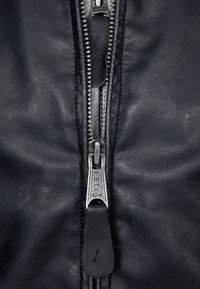 Sort læderjakke med en fremtrædende sølvfarvet lynlås med et "ALPHA" logo på zip-pull, glat tekstur og detaljeret syning langs sømmene.