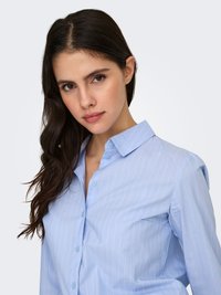 Young woman with long dark hair wearing a light blue pinstriped button-up shirt, looking directly with a neutral expression.