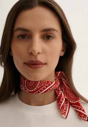 Czerwona chusta w stylu bandany z wzorem paisley i białymi akcentami, zawiązana na szyi, w połączeniu z prostą białą koszulką. Gładka tekstura.