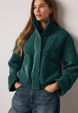 Quilted green bomber jacket with a high collar, elastic hem, and visible stitching patterns, paired with dark denim jeans.