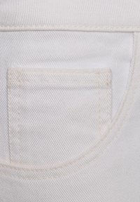 Detailní záběr bílé denimové látky s přišitým obdélníkovým kapsou a zakřivenými švovými liniemi.