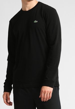 Homme portant un t-shirt noir uni à manches longues avec un petit logo crocodile vert sur la poitrine.