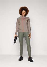Letvægts, tan farvet hættetrøje med rød lynlås, parret med olivengrønne leggings. Model holder en sort vandflaske og har sorte sneakers på.
