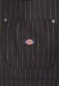 Crni traper pregač s bijelim prugama, sadrži džep na prsima s kružnim Dickies logotipom i srebrnim kopčanjem. Tekstuirana tkanina.