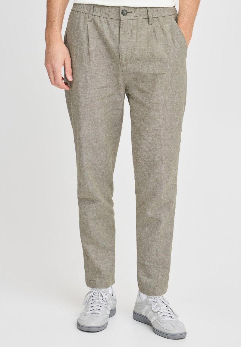 Man met lichtbruine linnen broek, wit overhemd en grijze sneakers, staat met handen in de zakken tegen een effen witte achtergrond.