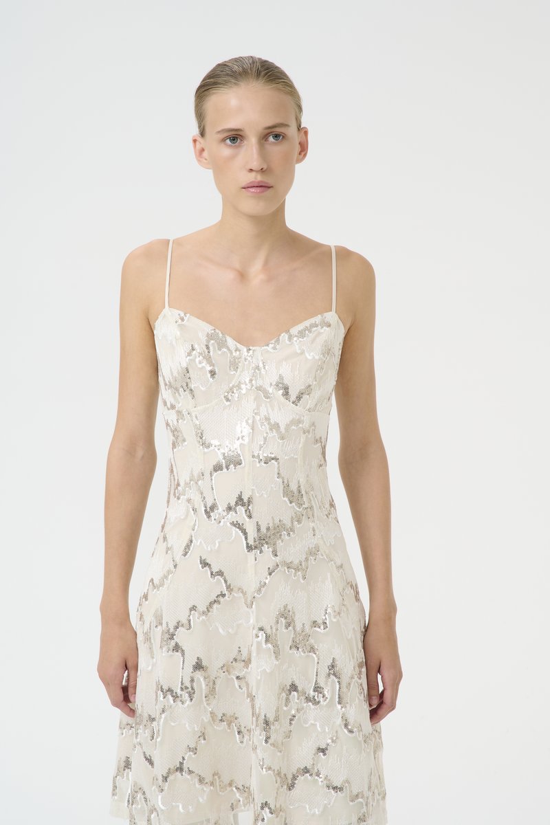 Valge kleit õhukeste rihmadega, millel on laineline hõbedane ja beež muster, valmistatud tekstuurses kangas, millel on liibuv kehaosa ja voolav seelik.