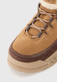 Botín de ante marrón con una suela de goma texturizada marrón, costuras en contraste y un diseño de cordones que presenta cordones con patrón y un collar acolchado.