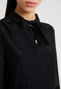 Zwarte blouse met een strikdetail bij de hals en een keyhole-halslijn. Zachte, gladde stof met een lichte glans en een losse pasvorm. Lange mouwen.