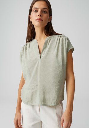 Femme portant un chemisier vert clair à manches courtes et col en V, et un pantalon blanc, debout devant un fond gris clair uni.