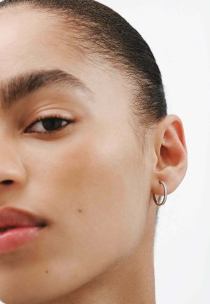 Pendiente de aro plateado en un modelo, con un diseño elegante y un acabado pulido que ofrece un sutil destello. Primer plano de la oreja y la cara.