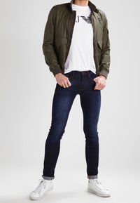 Olivgrön bomberjacka, vit grafisk t-shirt med logotyp, mörkblå smala jeans och vita sneakers. Slät textur med ribbade muddar.