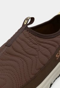 Rjava natikača z zaletno teksturo, z valovitim šivanjem in majhno zanko. Dodatki v svetlo rjavi barvi in smetanova podplat.