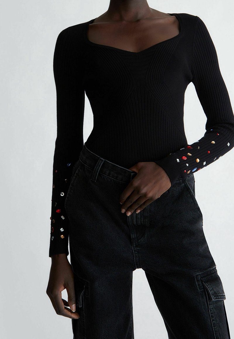 Femme portant un haut noir côtelé à manches longues avec des embellissements multicolores sur les manches et un pantalon cargo noir taille haute.