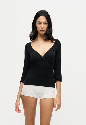 Donna con capelli scuri fino alle spalle indossa una maglia nera a maniche lunghe con scollo a V e bordo in pizzo e pantaloncini bianchi, in piedi davanti a uno sfondo chiaro e semplice.