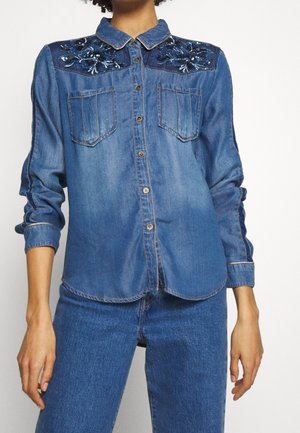 Femme portant une chemise en denim bleu moyen avec une broderie bleu foncé sur l'empiècement et un jean en denim bleu assorti.
