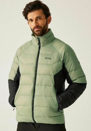 Mężczyzna w zielono-czarnej pikowanej kurtce z zamykanymi kieszeniami i logo "Regatta Great Outdoors" na piersi, stoi z rękami w kieszeniach.