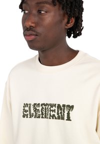 Cream-colored sweatshirt featuring a textured olive green "ELEMENT" graphic print with varying font styles and designs. Textured fabric.