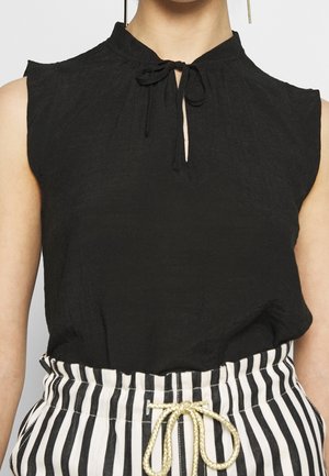 Mouwloze zwarte blouse met een klein sleutelgat en een striksluiting bij de hals, gecombineerd met hooggetailleerde zwart-witte gestreepte broek met een gouden trekkoord.