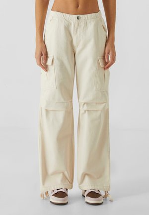 Pantalones cargo beige con cintura alta, que presentan grandes bolsillos laterales, un corte relajado y un detalle de cordón en el dobladillo. Combinados con zapatillas.