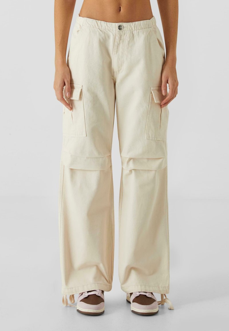 Beige cargo bukser med høj talje, der har store sidelommer, en afslappet pasform og detaljer med snøre ved kanten. Båret med sneakers.