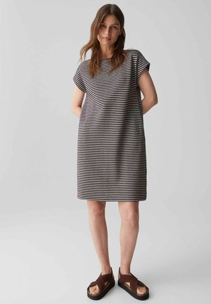 Frau steht mit den Händen hinter dem Rücken, trägt ein kurzärmeliges, knielanges schwarz-weiß gestreiftes Kleid und braune Plateausandalen.