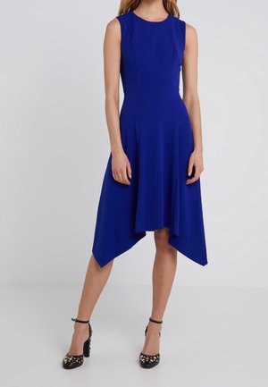 Mujer con vestido sin mangas azul real con dobladillo asimétrico y tacones altos negros con tachuelas doradas, posando sobre un fondo claro y liso.