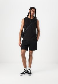Fekete ujjatlan pamut tank top, mellzsebbel, fekete hálós rövidnadrággal kiegészítve. A lábbeli fekete-fehér sportcipő.