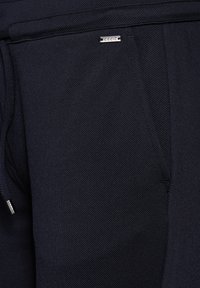 Marineblauwe textuurbroek met een ritszak en een klein metalen logo-tag. Gemaakt van een duurzame, ademende stof met een gladde afwerking.