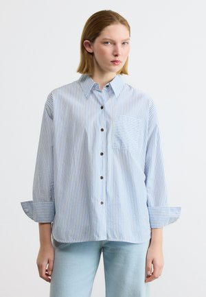 Junge Frau trägt ein lockeres, hellblau-weiß gestreiftes Hemd mit aufgekrempelten Ärmeln und hellblaue Jeans, steht still.