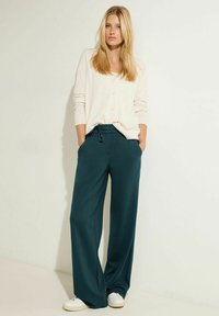 Květinové kalhoty v barvě teal se širokými nohavicemi, elastickým pasem a bočními kapsami, v kombinaci s světlým béžovým kardiganem na knoflíky a bílými teniskami.