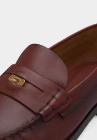 Καφέ δερμάτινος μοκασίνι με χρυσό έμβλημα και διακοσμητική ραφή στο επάνω μέρος του παπουτσιού.