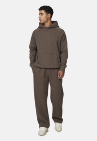 Giovane in felpa marrone e pantaloni abbinati, mani in tasca, guarda di lato, indossa sneaker bianche.