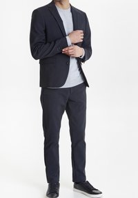 Donkerblauwe colbert over een lichtgrijs vest, gecombineerd met bijpassende marineblauwe broek en zwarte sneakers. Glad weefsel met een getailleerde pasvorm.