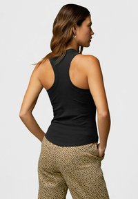 Schwarzes ärmelloses Tank-Top mit geripptem Muster, enganliegendem Design und weiten Armausschnitten, kombiniert mit beigen Hosen, die ein schwarzes Punktmuster aufweisen.