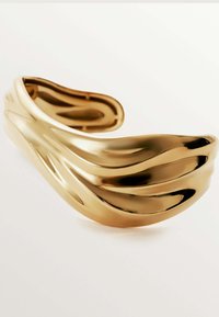 Pulsera de puño en tono dorado que presenta un diseño ondulado con acabado pulido y textura en capas. Su forma abierta permite un ajuste personalizado.