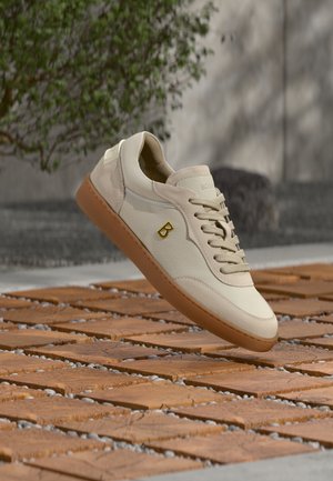 Pantof sport bej din piele cu talpă maro și logo „B” auriu, suspendat deasupra unei suprafețe cu textură de lemn, cu verdeață în fundal.