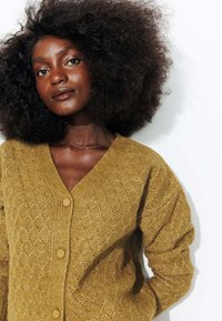 Cardigan en tricot jaune moutarde avec un col en V, motif texturé, manches longues et fermeture à boutons. Matière douce avec une apparence cozy.