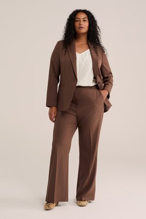 Femme aux cheveux bouclés portant un tailleur marron, un haut blanc et des chaussures beige, debout avec une main dans la poche devant un fond uni.
