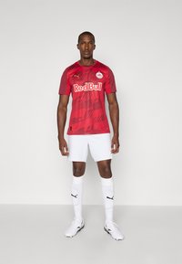 Maillot de football rouge avec des motifs graphiques, shorts rouges, chaussettes blanches et crampons. Arbore le logo Red Bull et la marque Puma sur le maillot.