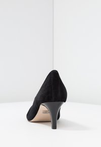 Chaussure à talon haut en suede noir avec un bout pointu et un talon fin texturé. La semelle est nude et il y a un détail de marque discret près du talon.