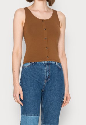 Kobieta w brązowym bezrękawnikowym, zapinanym na guziki krótkim topie i dżinsach dwuodcieniowych niebieskich, stojąca na jednolitym jasnym tle.