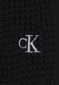 Črna pletena kapa z vtkano teksturo, ki ima izstopajoč bel izvezen logotip 'cK'.