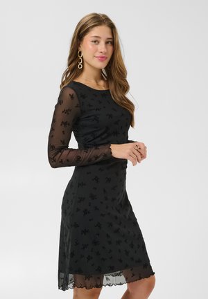 Une femme porte une robe noire longueur genou, avec des manches et un ourlet transparents à motifs floraux. Elle porte des boucles d’oreilles et des bagues en or, sur un fond clair et uni.