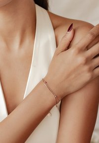 La pulsera de oro rosa presenta un diseño de cadena delgada con un acento curvado y un pequeño charm, exhibida en un brazo desnudo sobre un fondo claro.