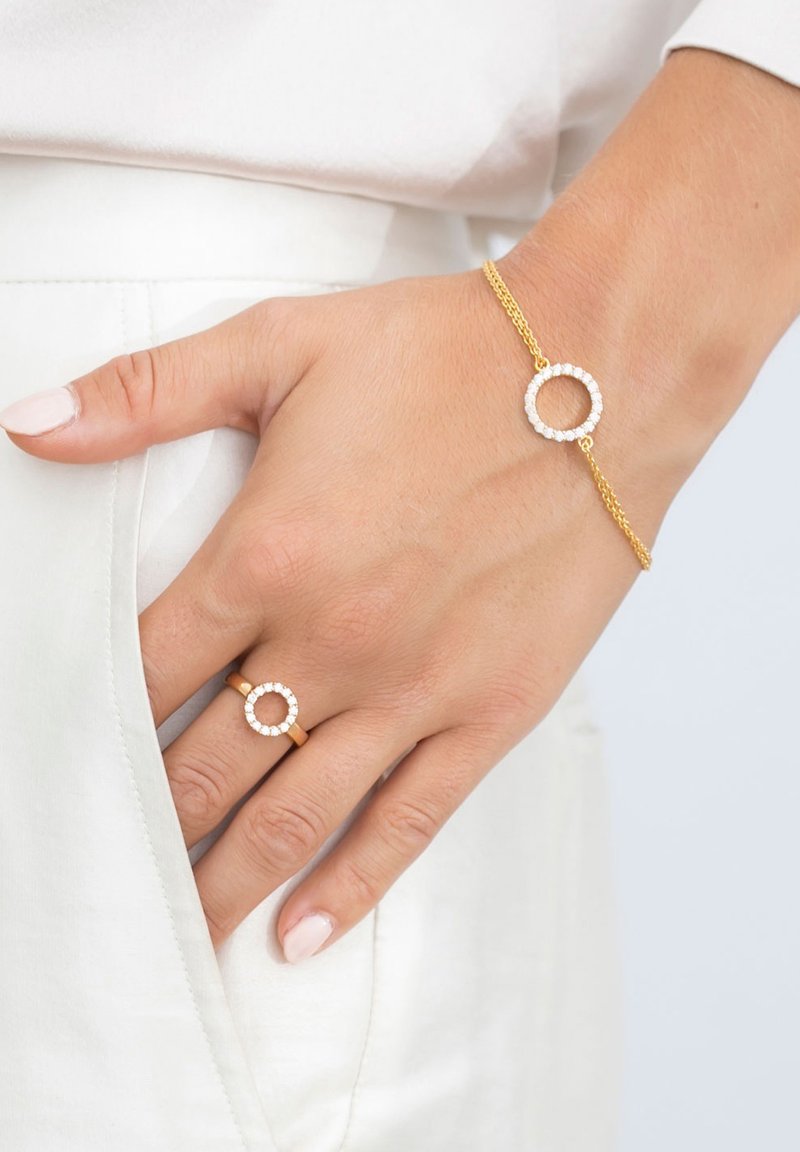 Guldarmband och ring med cirkulära mönster som har inlagda klara stenar. Armbandet har en elegant kedja, och båda föremålen har en slät yta.