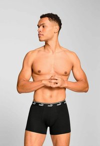 Muscular man with curly hair wearing black Puma boxer briefs, standing against a plain light gray background, looking to his left.