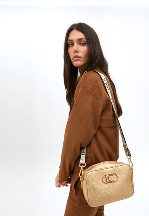 Une femme aux longs cheveux noirs fait face à la caméra, portant une tenue en daim marron et portant un petit sac bandoulière doré.