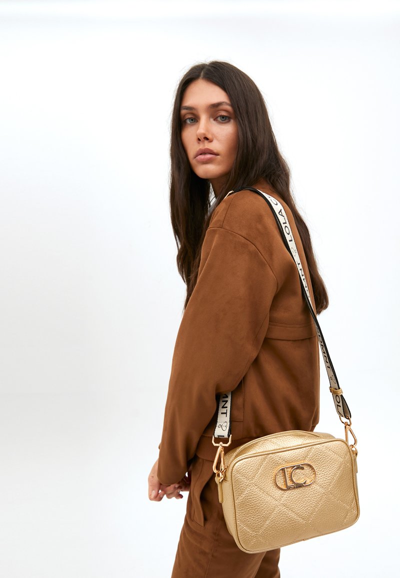 Una donna con lunghi capelli scuri guarda verso la telecamera, indossando un outfit in suede marrone e portando una piccola borsa a tracolla dorata.